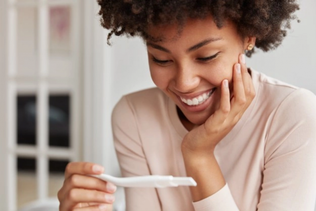 Mulher sorri ao conferir teste de gravidez: mesmo na primeira semana após a concepção, os indícios da gravidez já podem aparecer. Por isso, conheça os primeiros sinais e sintomas da gravidez!