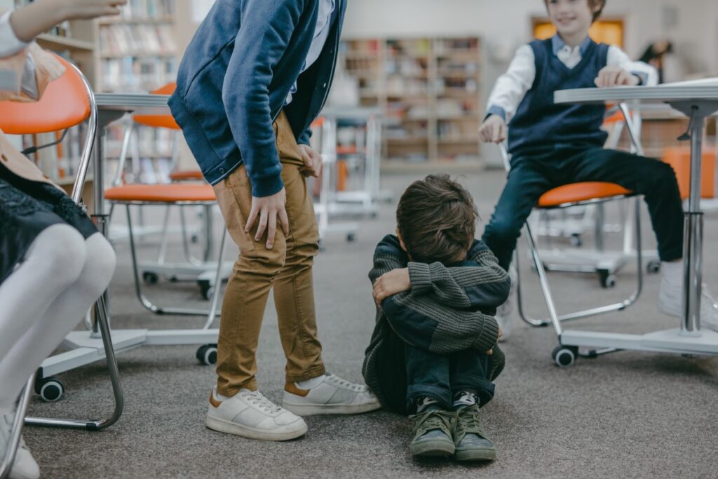 Crianças riem de um menino sentado no chão e com a cara escondida entre os braços por ter vergonha: conheça 5 histórias para falar de bullying com crianças
