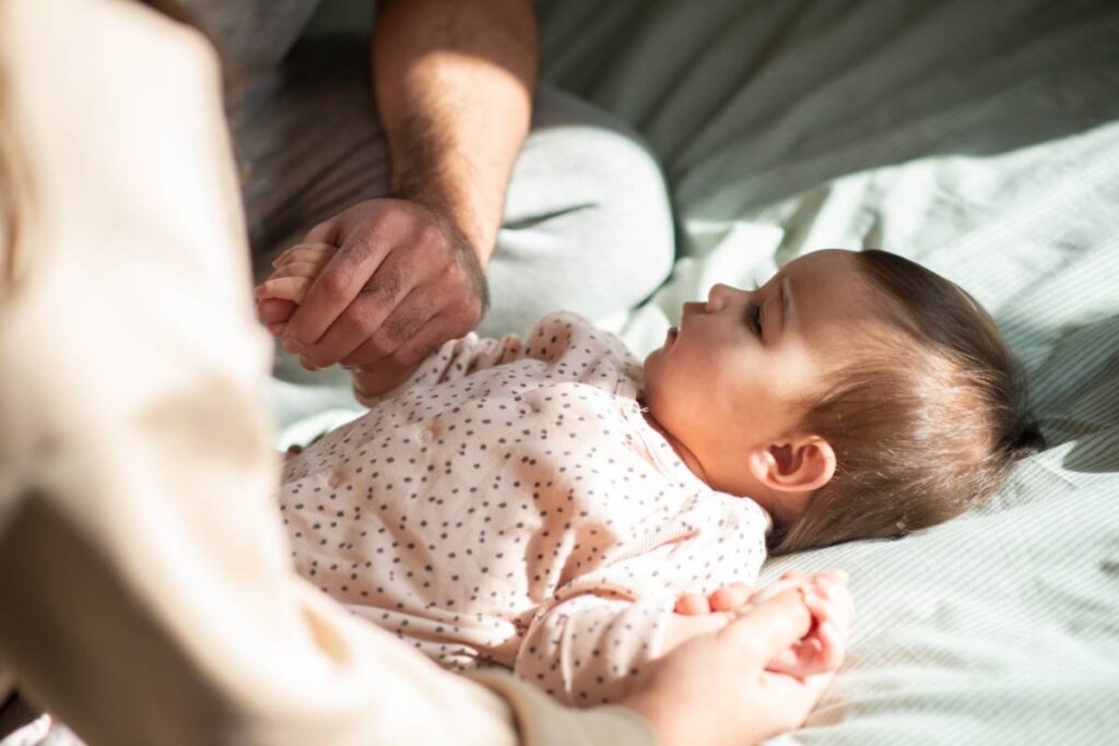 Pai e mão seguram a mão da sua bebê deitada na cama: logo nos primeiros dias de vida, é necessário que a nenê passe por uma série de exames, inclusive alguns antes mesmo de deixar a maternidade
